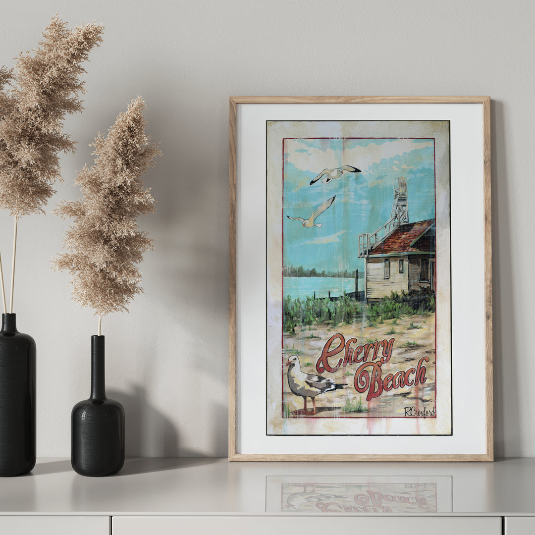 Cherry Beach Poster by Rob Croxford in a wooden frame on a drawer with black mantel bottle