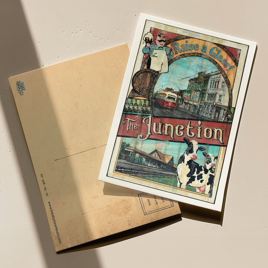 The Junction - Raise a Glass Toronto Postcard by Rob Croxford with front and backend of postcard visible on a table